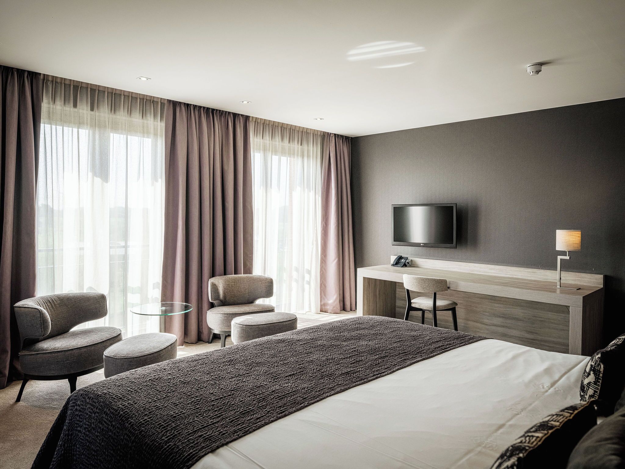Een moderne hotelkamer met een groot bed, een bureau met stoel, een aan de muur bevestigde televisie en een bijzettafeltje met lamp.