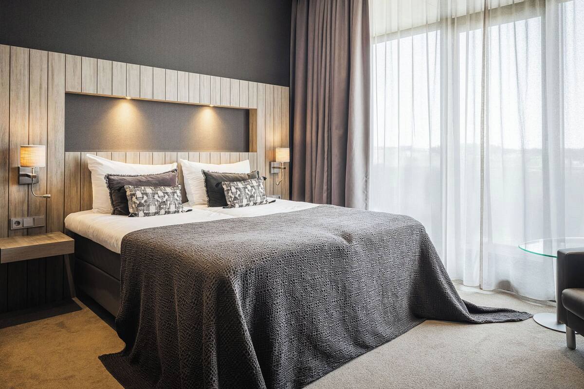 Een moderne hotelkamer met een groot bed, twee nachtkastjes met lampen, een donker hoofdeinde en een groot raam met dunne gordijnen.