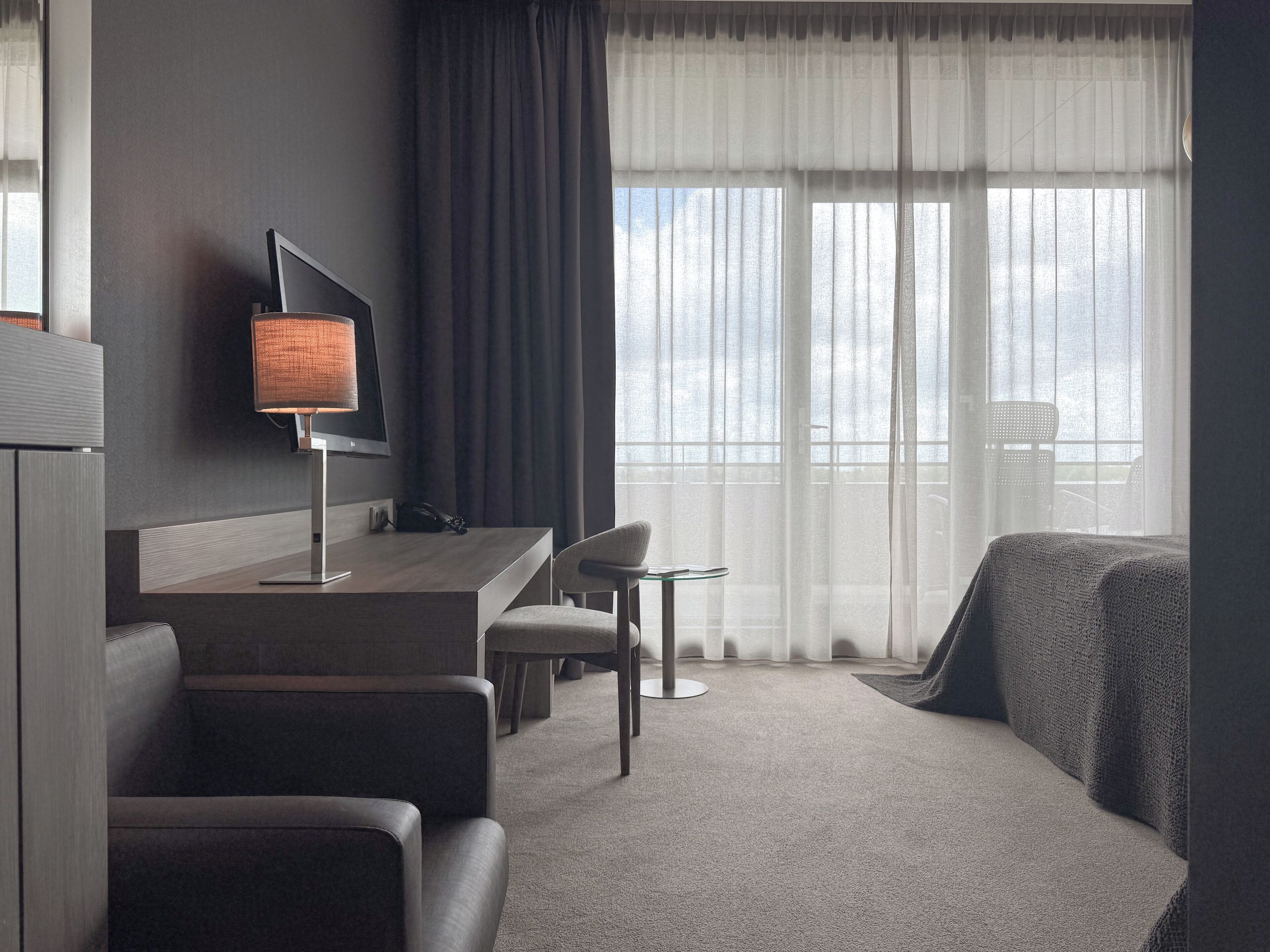 A modern hotel room with a dark-colored sofa, a small round table, a desk with a lamp, and a large window with sheer curtains.