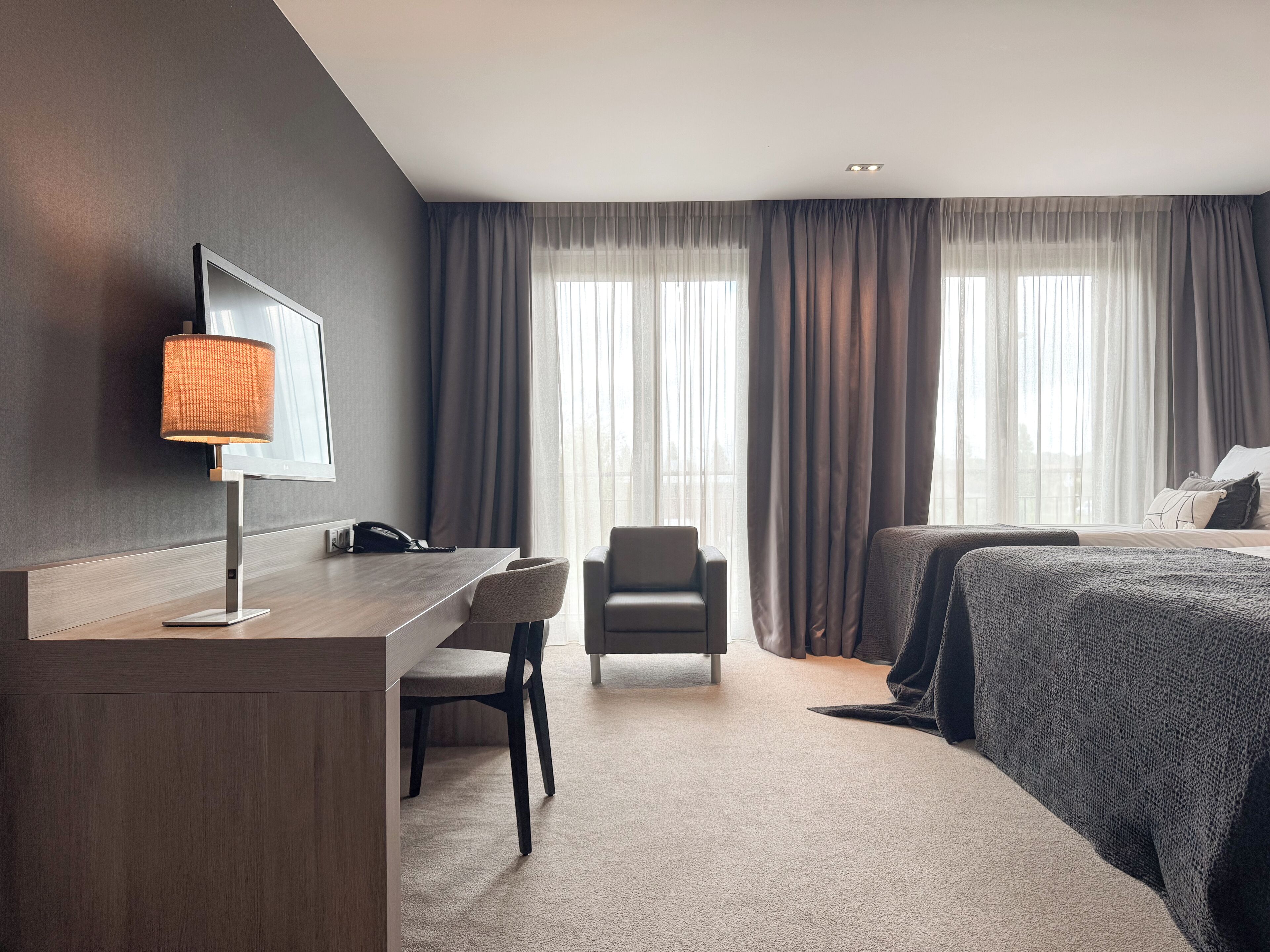 A hotel room with a desk, chair, bed, and large windows with curtains.