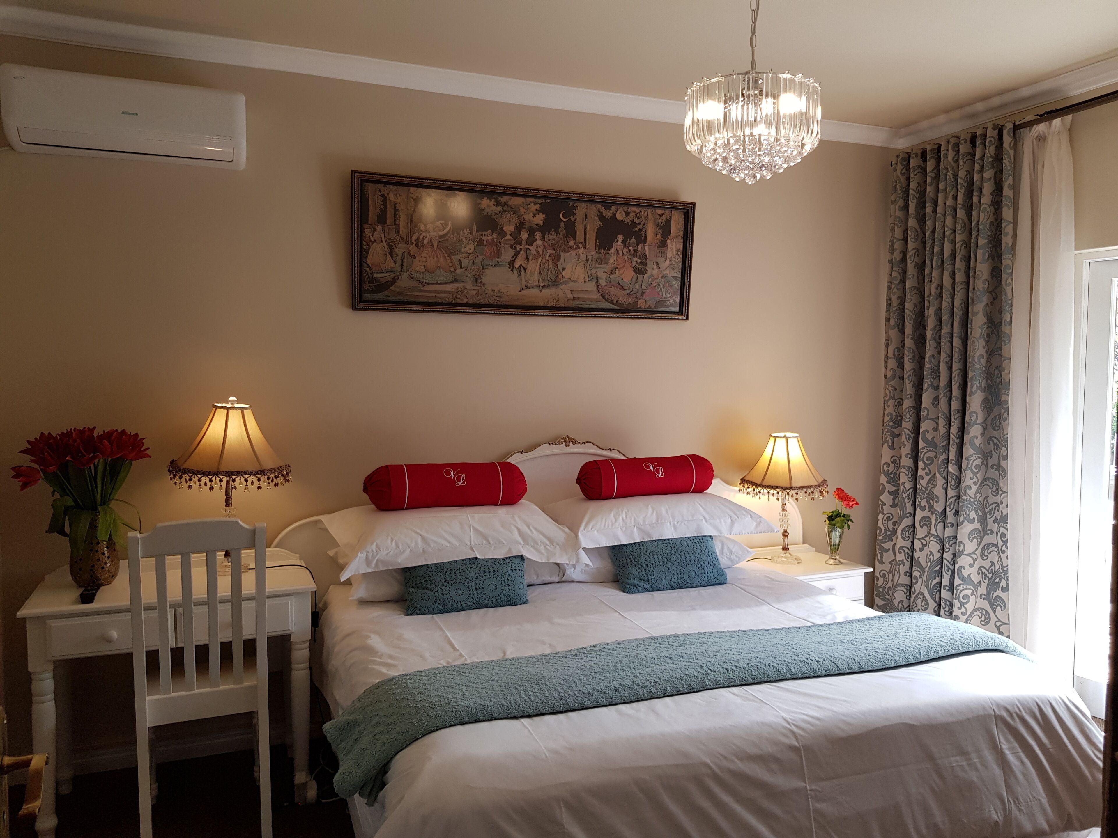 Une chambre d’hôtel avec un lit, deux tables de chevet, une chaise, un lustre, un tableau et une fenêtre avec des rideaux.