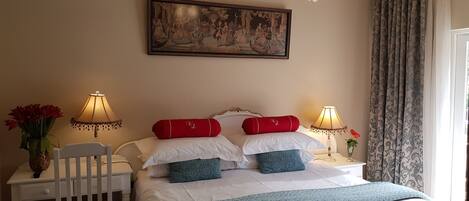 Une chambre d’hôtel avec un lit, deux tables de chevet, une chaise, un lustre, un tableau et une fenêtre avec des rideaux.