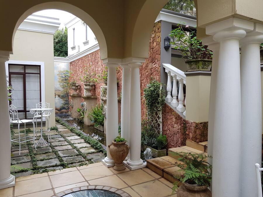 Een binnentuin met een stenen pad, witte zuilen, een klein fonteintje en potplanten.