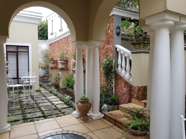 A courtyard with a stone pathway, white columns, a small fountain, and potted plants. - Villa Lugano (Johannesburg South)