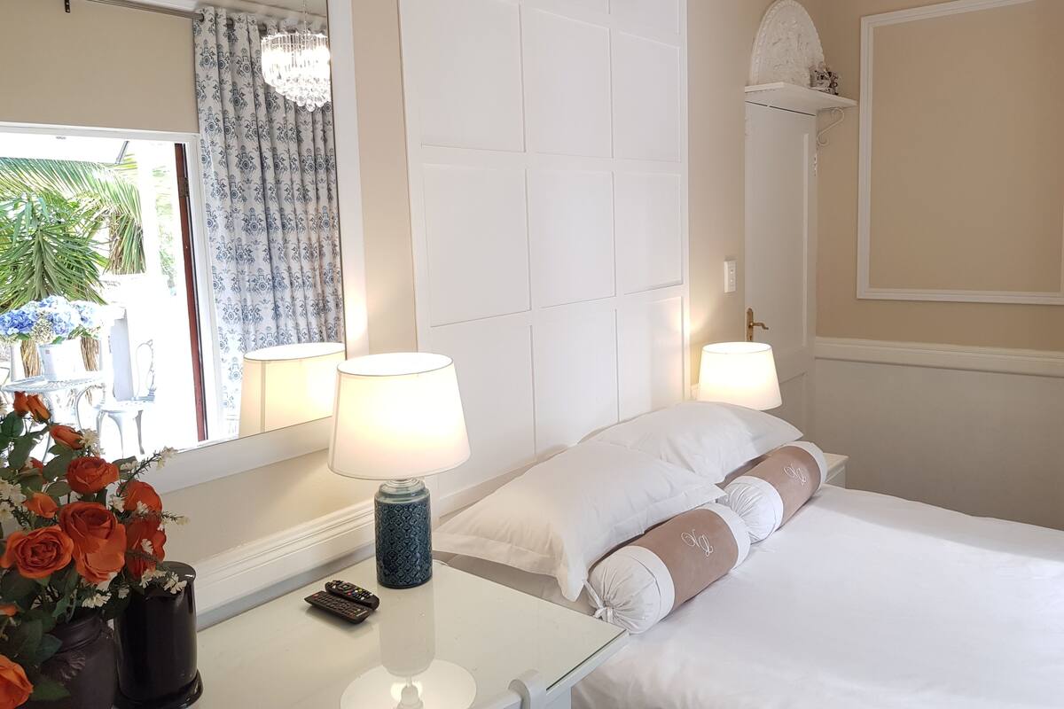 A hotel room with a bed, two bedside lamps, a nightstand with a remote control, a vase with flowers, and a window with curtains.