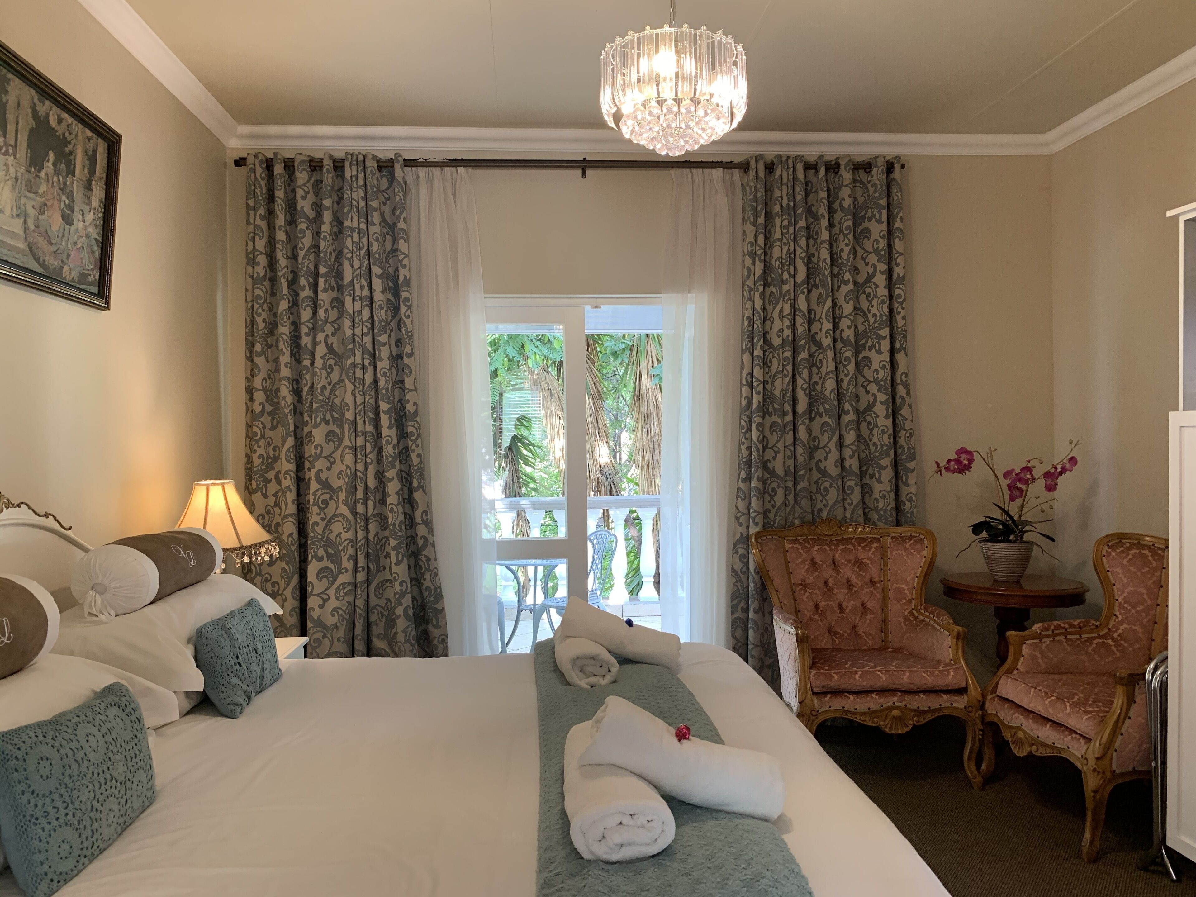A hotel room with a large bed, a chandelier, a balcony with curtains, and a seating area with chairs.