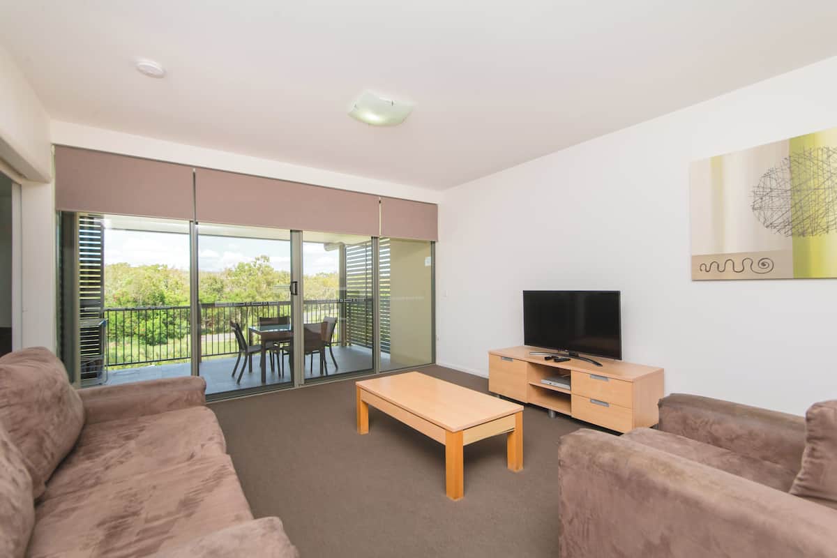 Ein modernes Wohnzimmer mit einer Couch, einem Couchtisch, einem Fernseher und einer Glastür, die auf einen Balkon mit Tisch und Stühlen führt.