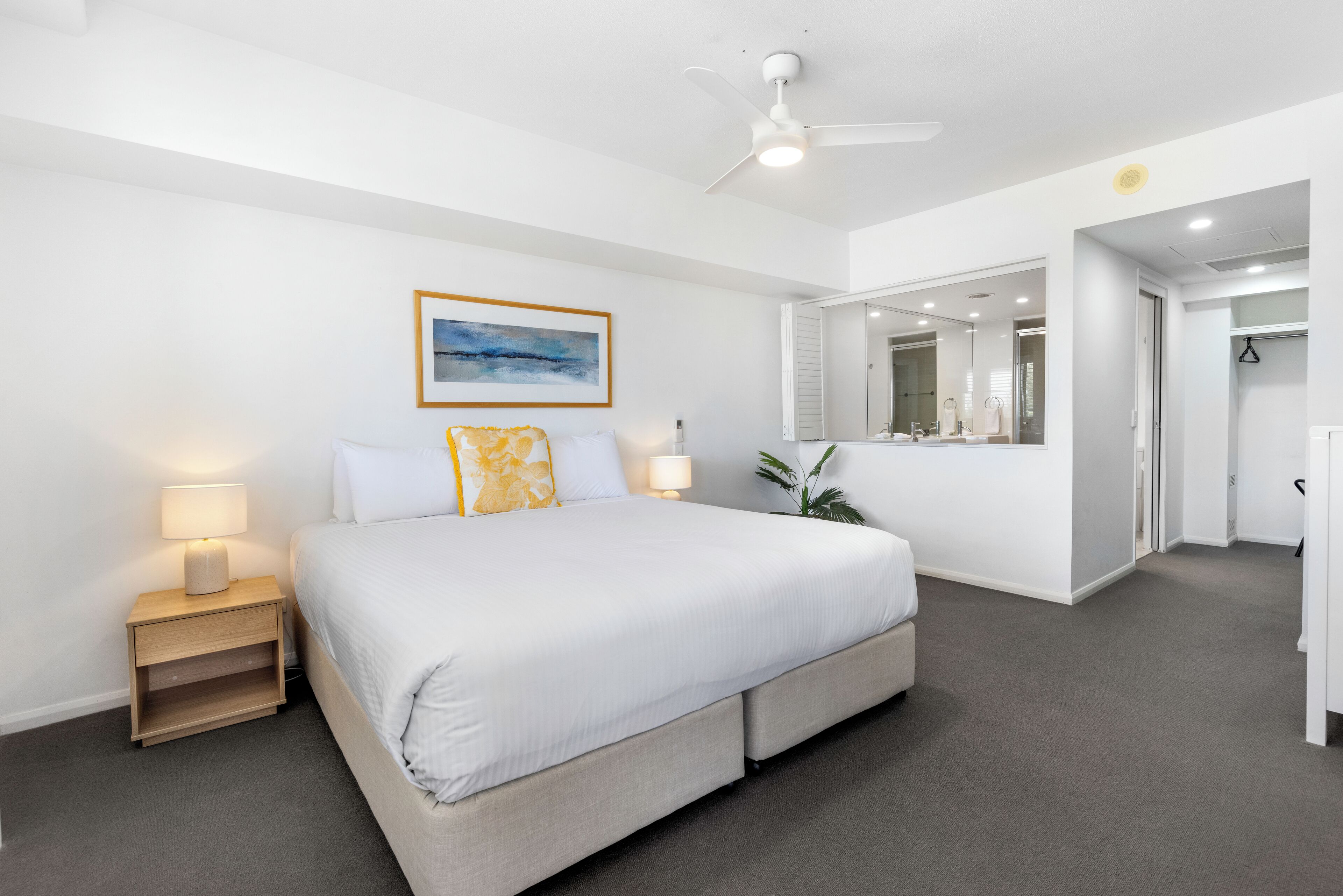 A modern bedroom with a large bed, bedside lamps, a ceiling fan, and a painting on the wall.
