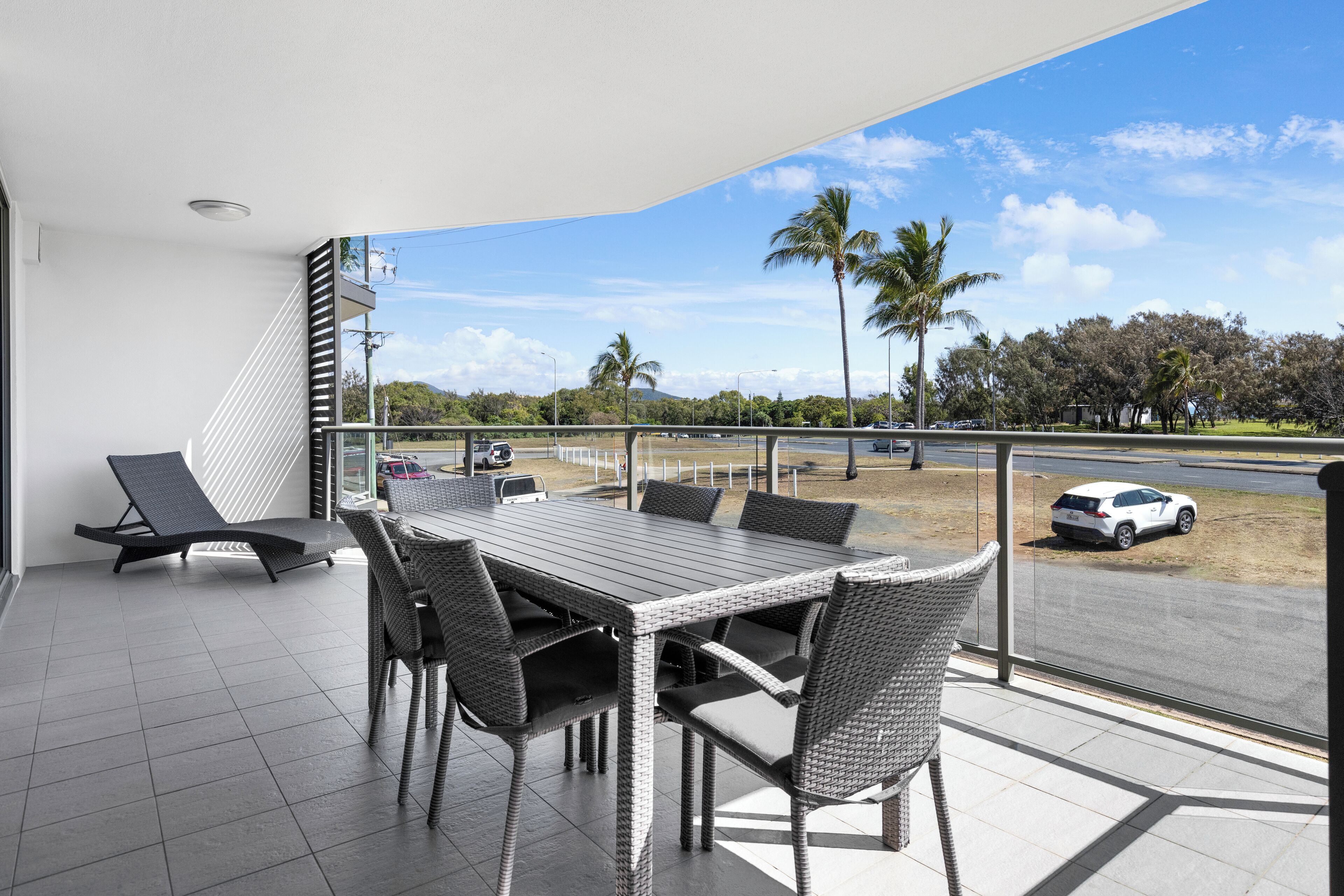 A balcony with a dining table and chairs, overlooking a road with cars and palm trees.