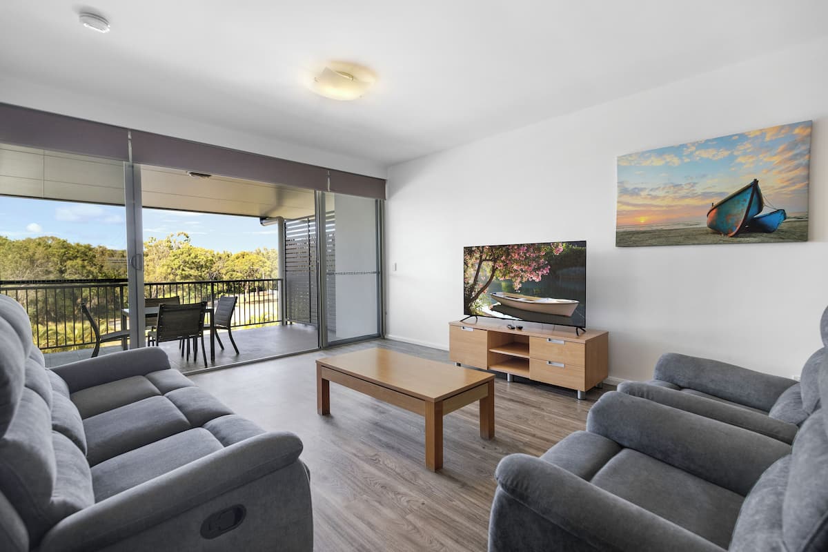 Ein modernes Wohnzimmer mit Sofa, Couchtisch und Fernseher auf einem Holzboden. Ein Balkon bietet Blick auf Bäume und einen Sonnenuntergang.
