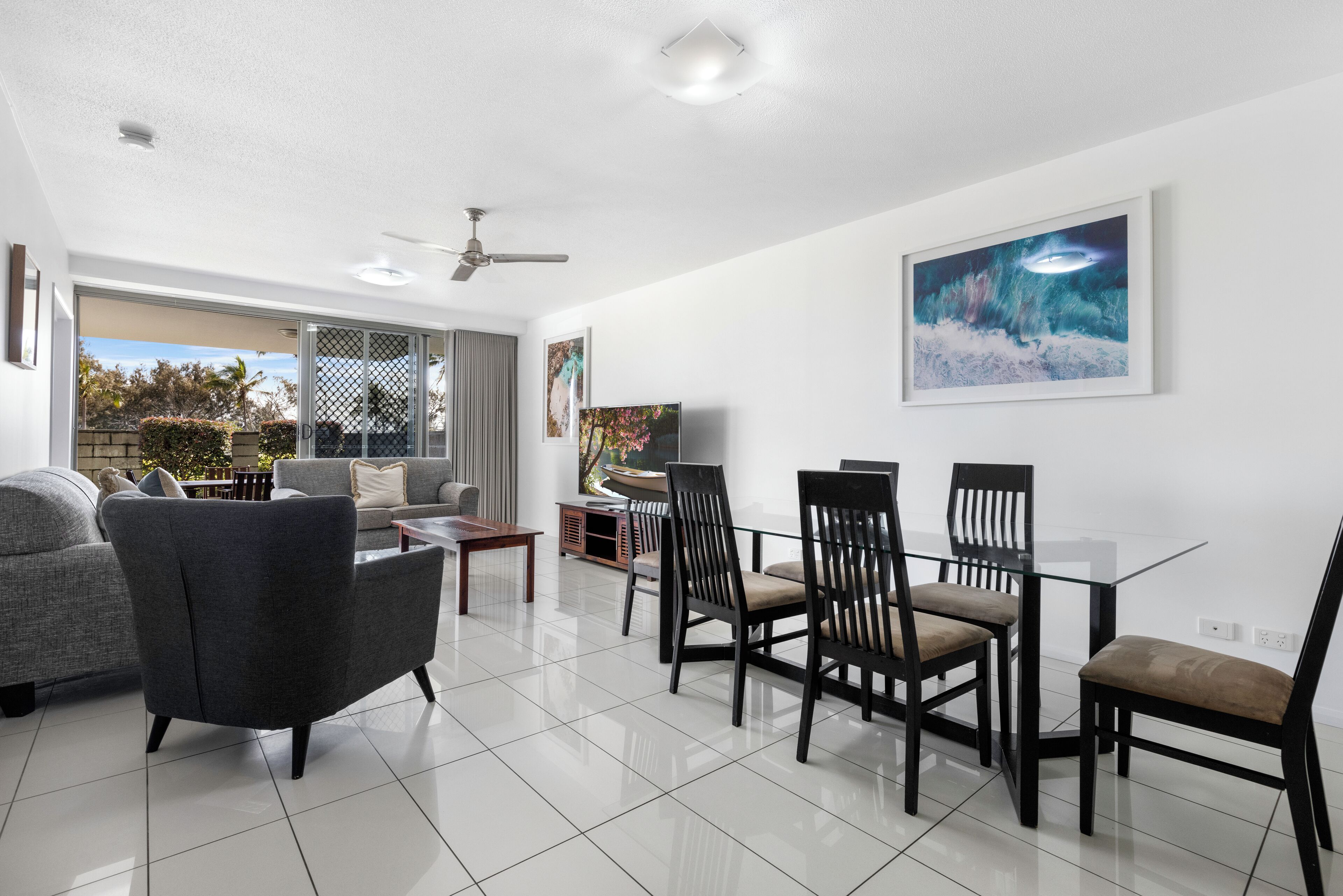 A modern living room with a glass dining table, dark chairs, a grey sofa, and a ceiling fan.