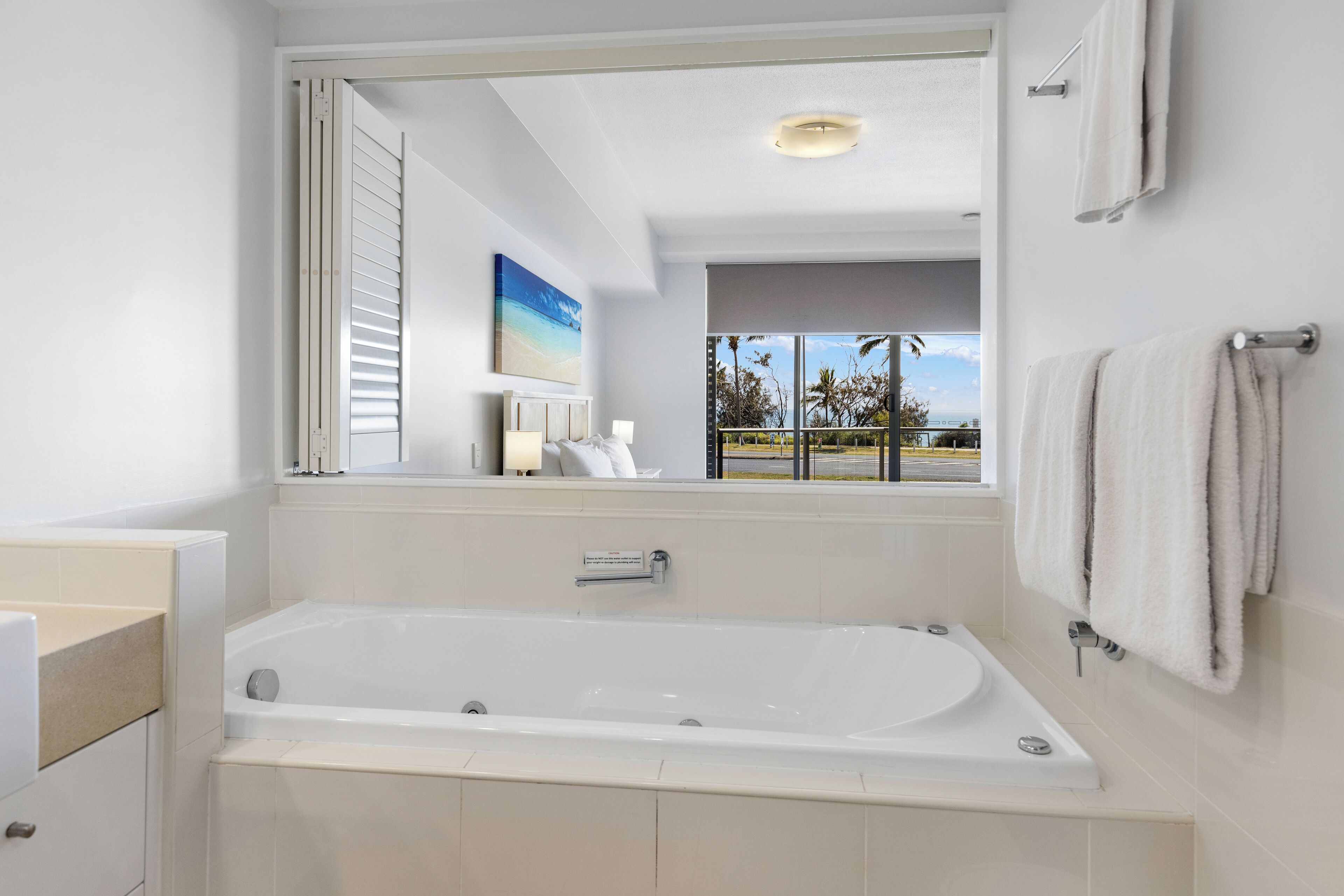 Ein modernes Badezimmer mit einer großen Badewanne, einem Fenster mit Blick auf Palmen und einem Handtuchhalter.