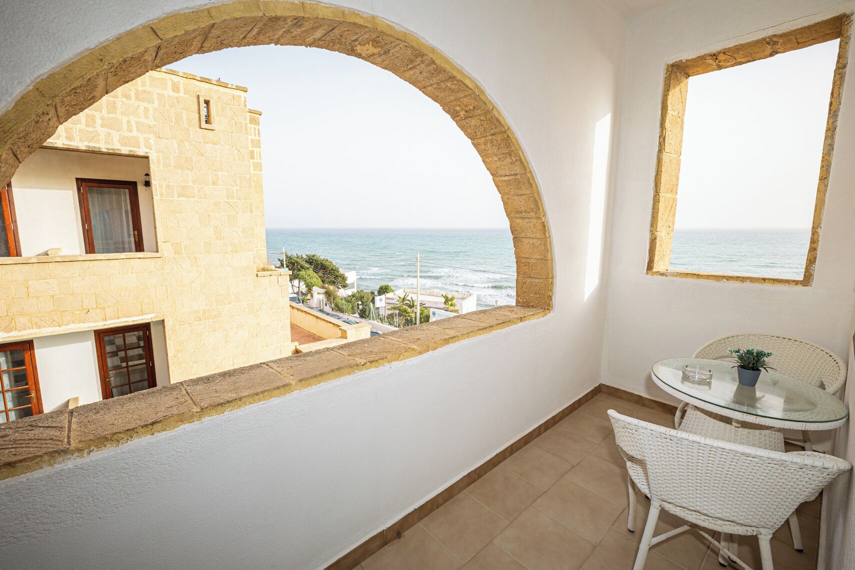 A balcony with a view of the sea, a small table with chairs, and a potted plant.