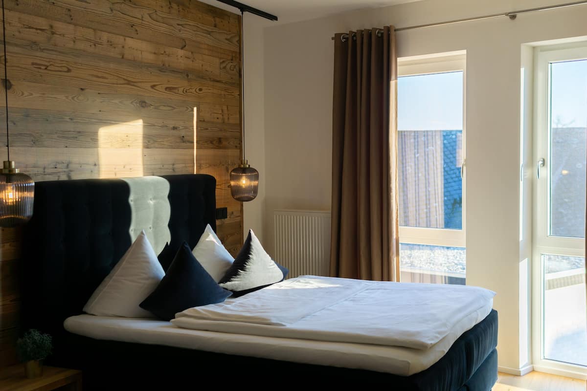 A bedroom with a large bed, wooden wall paneling, a window with curtains, and a small plant on a nightstand.