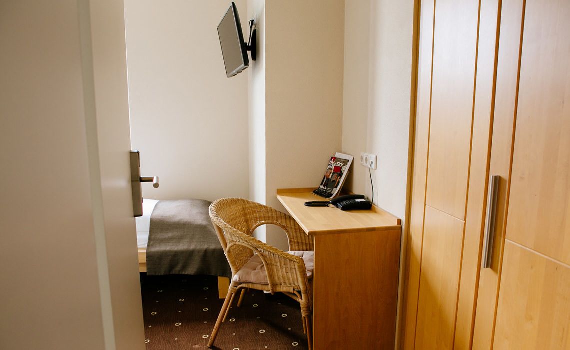 A small room with a bed, a wicker chair, a desk with a phone, and a magazine on the wall.