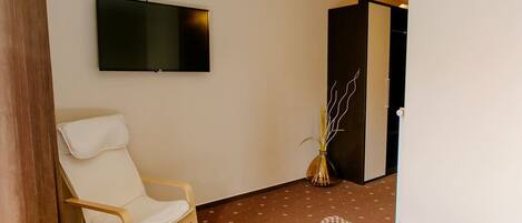 A hotel room with a flat-screen TV mounted on the wall, a wooden chair, a patterned rug, and a vase with branches.