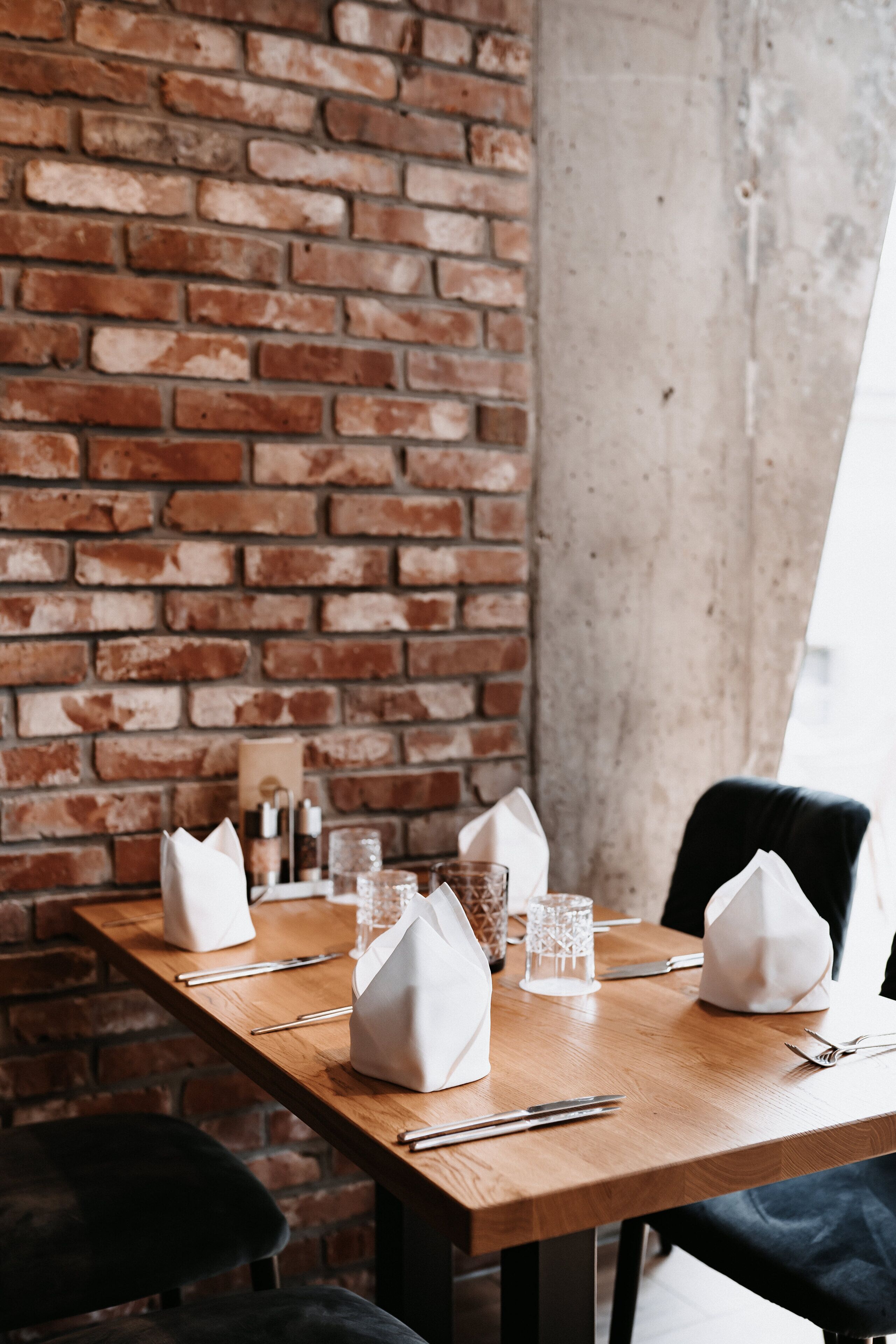 A dining table set with white napkins, cutlery, and glassware against a brick wall.