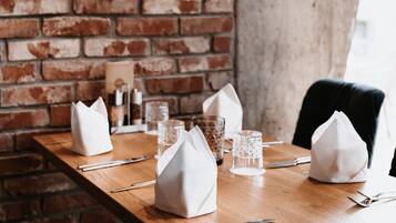 A dining table set with white napkins, cutlery, and glassware against a brick wall.