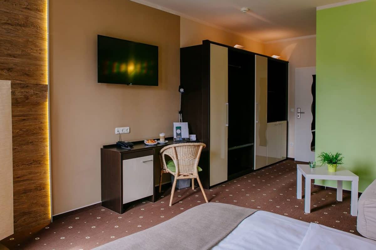 A hotel room with a bed, a desk with a chair, a television, a wardrobe, and a small table with a plant.