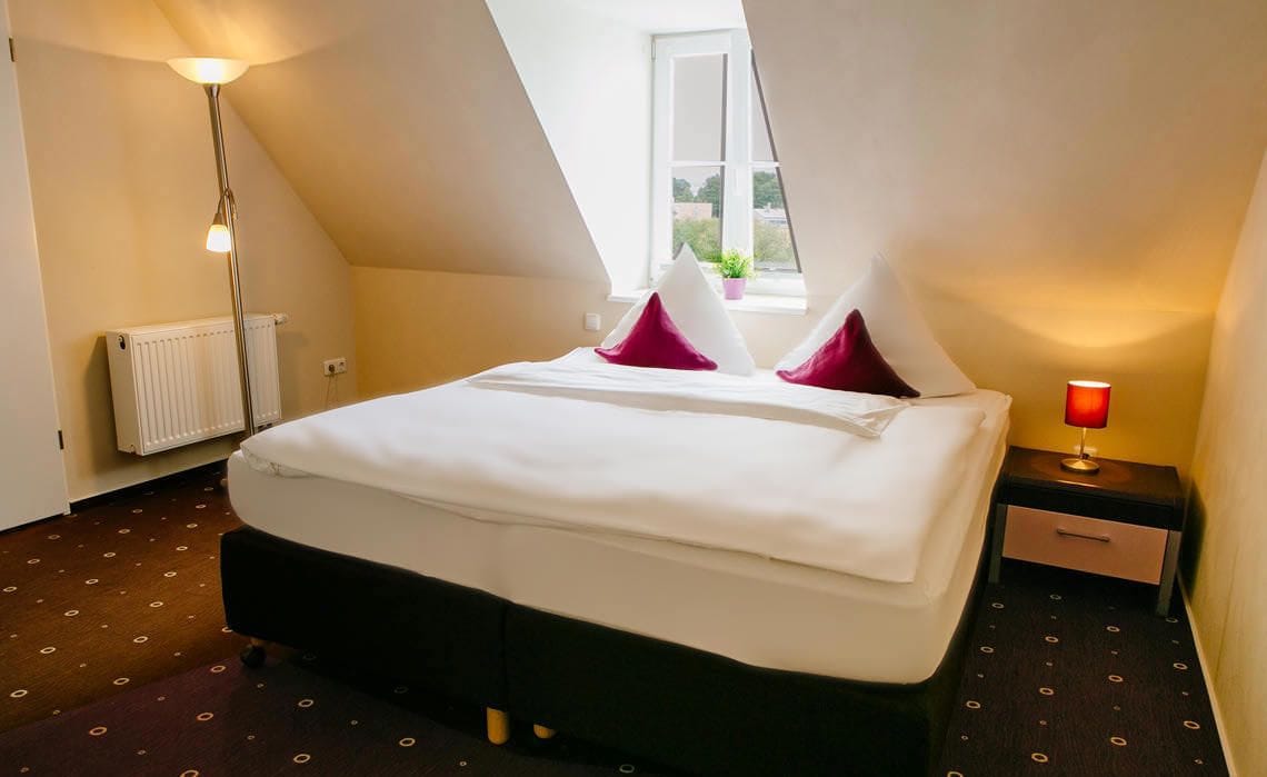 Un dormitorio con cama, mesita de noche, lámpara, radiador y ventana con planta.