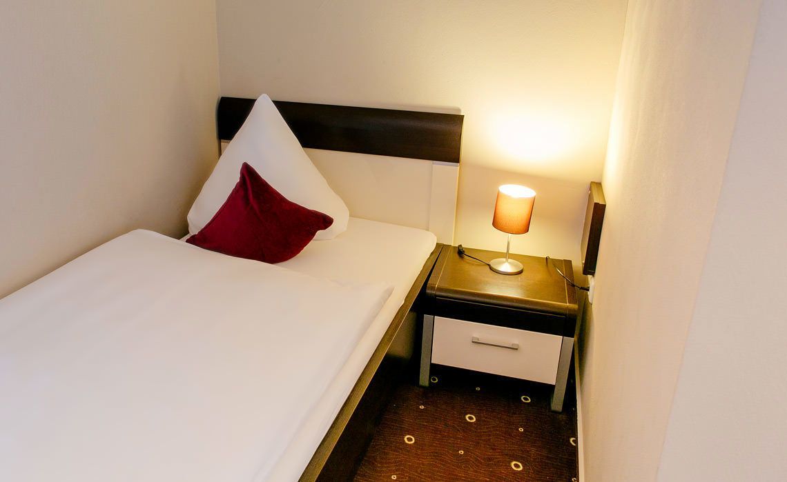 Un lit bien fait, avec des draps blancs et un oreiller rouge, une table de chevet avec une lampe et un petit tiroir.