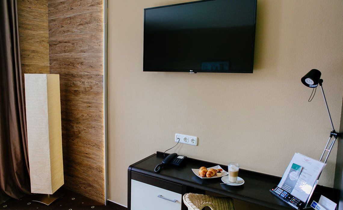 A hotel room with a flat-screen TV mounted on the wall, a desk with a phone, a laptop, a plate of croissants, a glass of coffee, and a lamp.