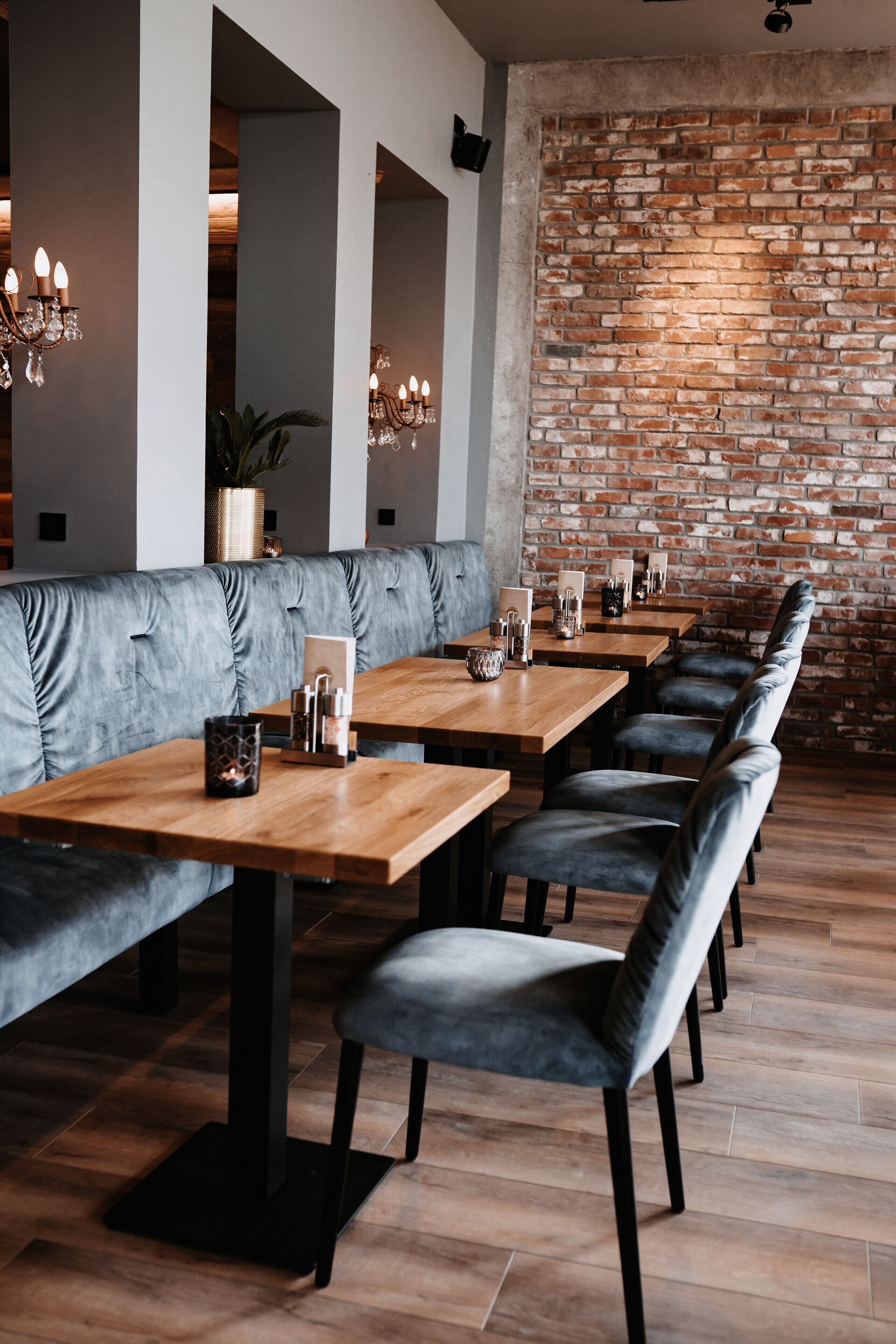A restaurant with a brick wall, grey upholstered booths, wooden tables, and black chairs.