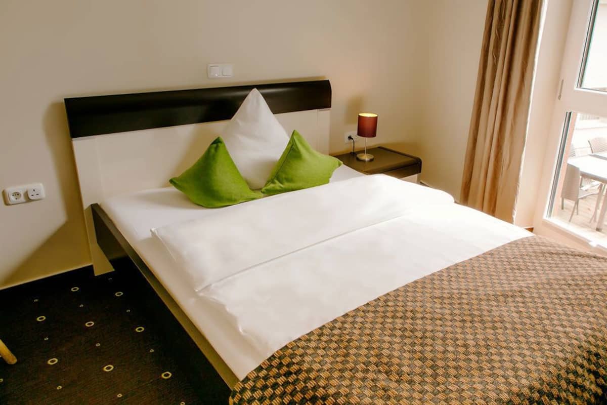 A hotel room with a bed, green and white pillows, a bedside lamp, and a view of the outside.