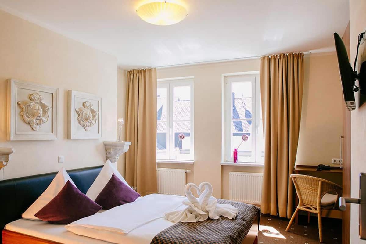 A hotel room with a bed, two wall-mounted artworks, a desk with a chair, and a window with curtains.