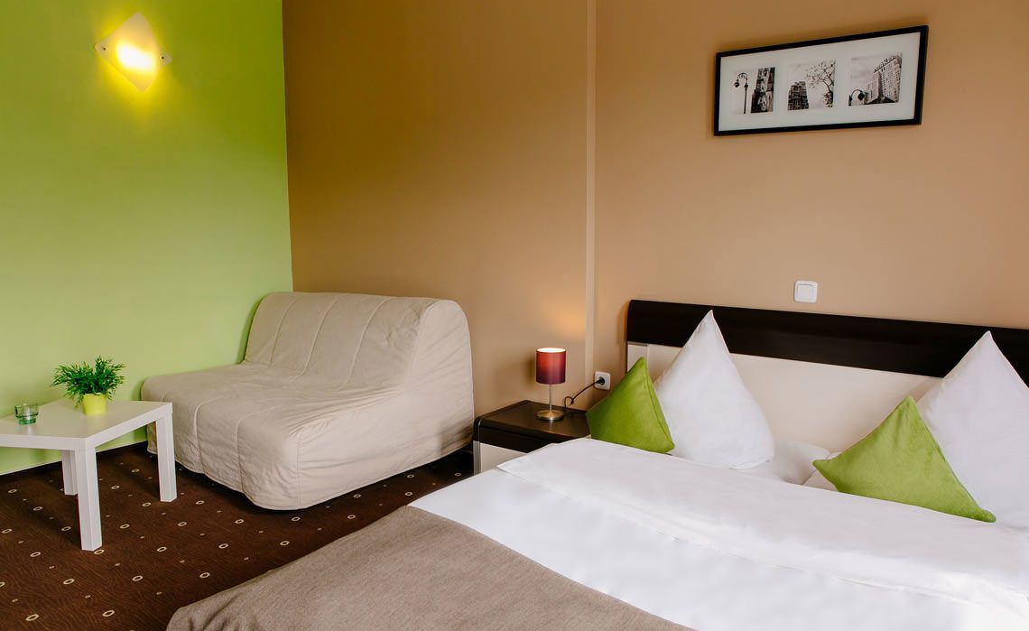 A hotel room with a bed, a chair, a nightstand with a lamp, a small table with a plant, and a wall-mounted picture.