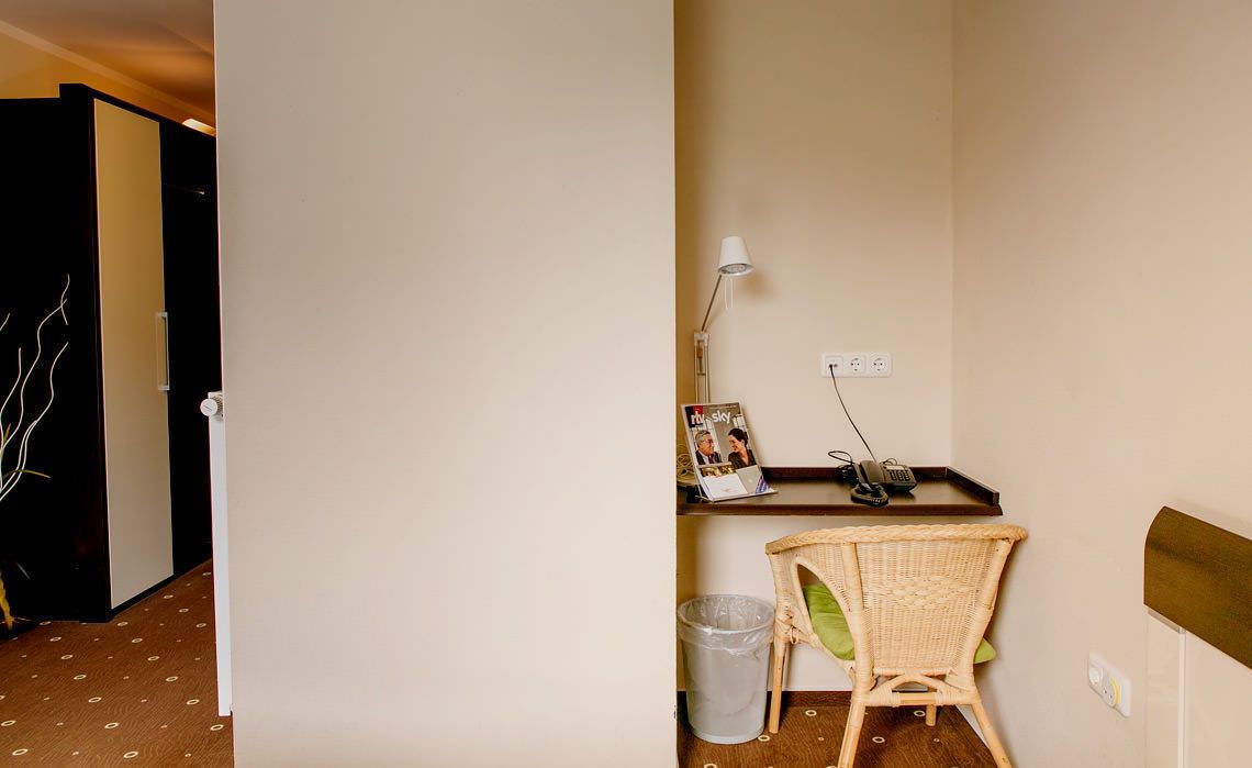 A hotel room with a wooden floor, a wicker chair, a small table with a lamp, a picture frame, and a trash bin.