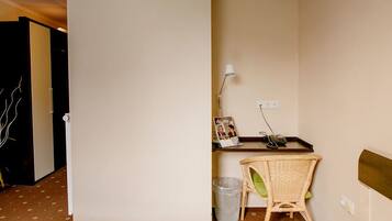 A hotel room with a wooden floor, a wicker chair, a small table with a lamp, a picture frame, and a trash bin.