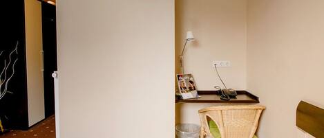 A hotel room with a wooden floor, a wicker chair, a small table with a lamp, a picture frame, and a trash bin.