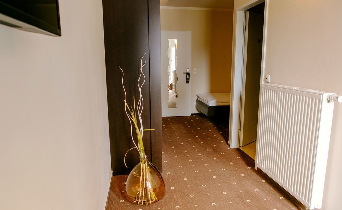 A hallway with a vase of branches, a radiator, and a bed in the distance.