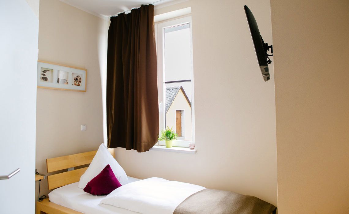 Een kleine kamer met een enkel bed, een raam, een tv aan de muur en een ingelijst schilderij.