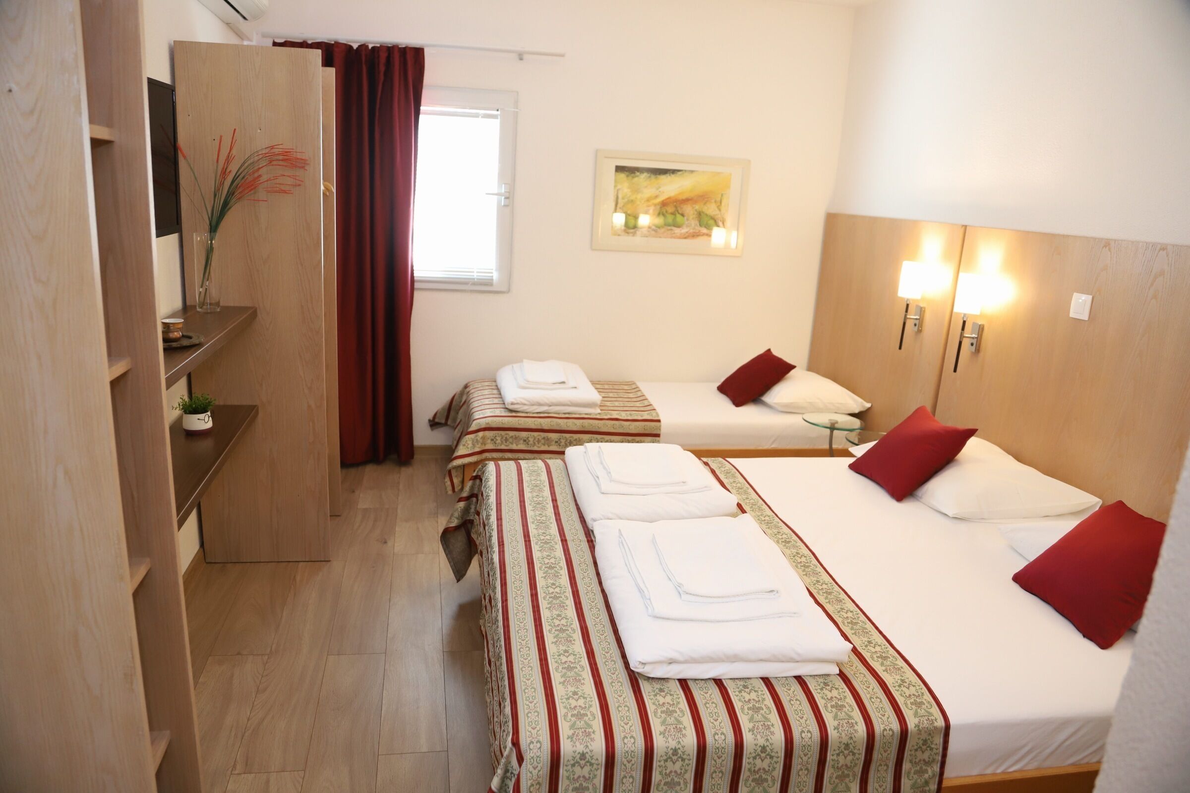 A hotel room with two single beds, a small desk, a mirror, a painting on the wall, and a window with curtains.