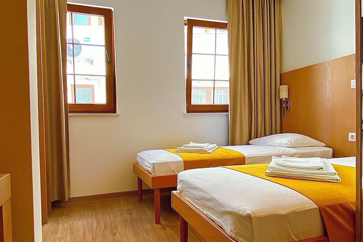 Habitación de hotel con dos camas, suelo de madera y dos ventanas con cortinas.