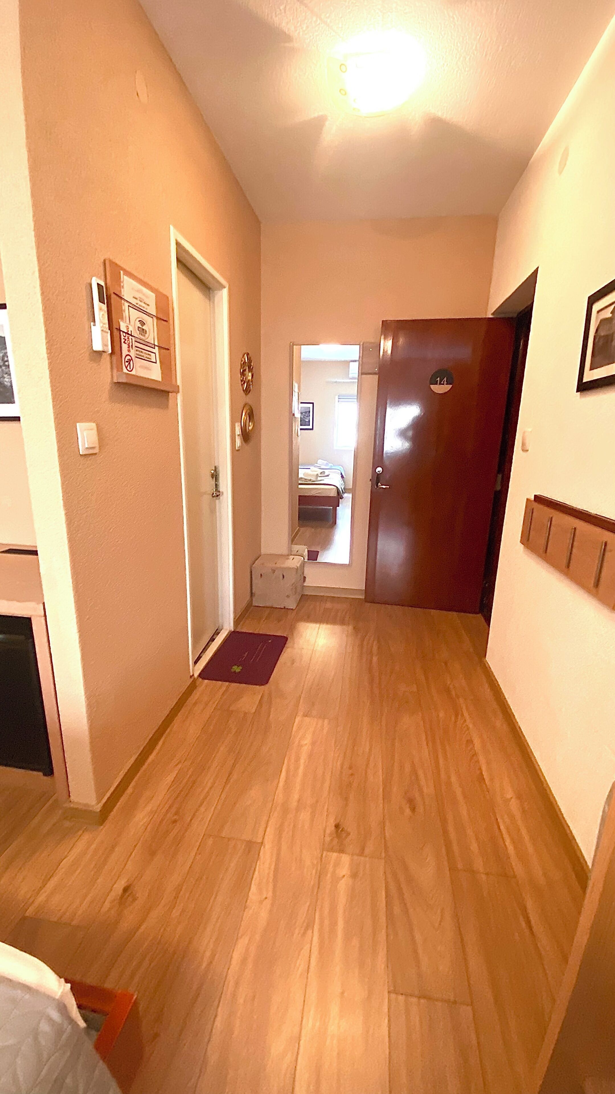A hallway with wooden flooring, a door with a number sign, a small mat, and a wall-mounted light fixture.