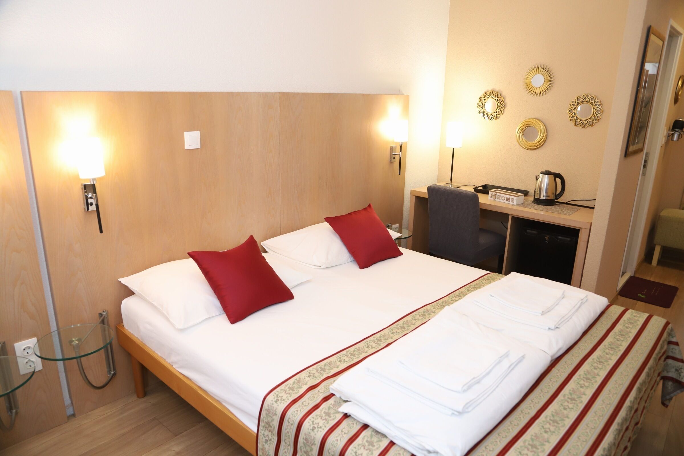 A hotel room with a wooden headboard, a bed with white linens and red pillows, a desk with a kettle, and wall decor.
