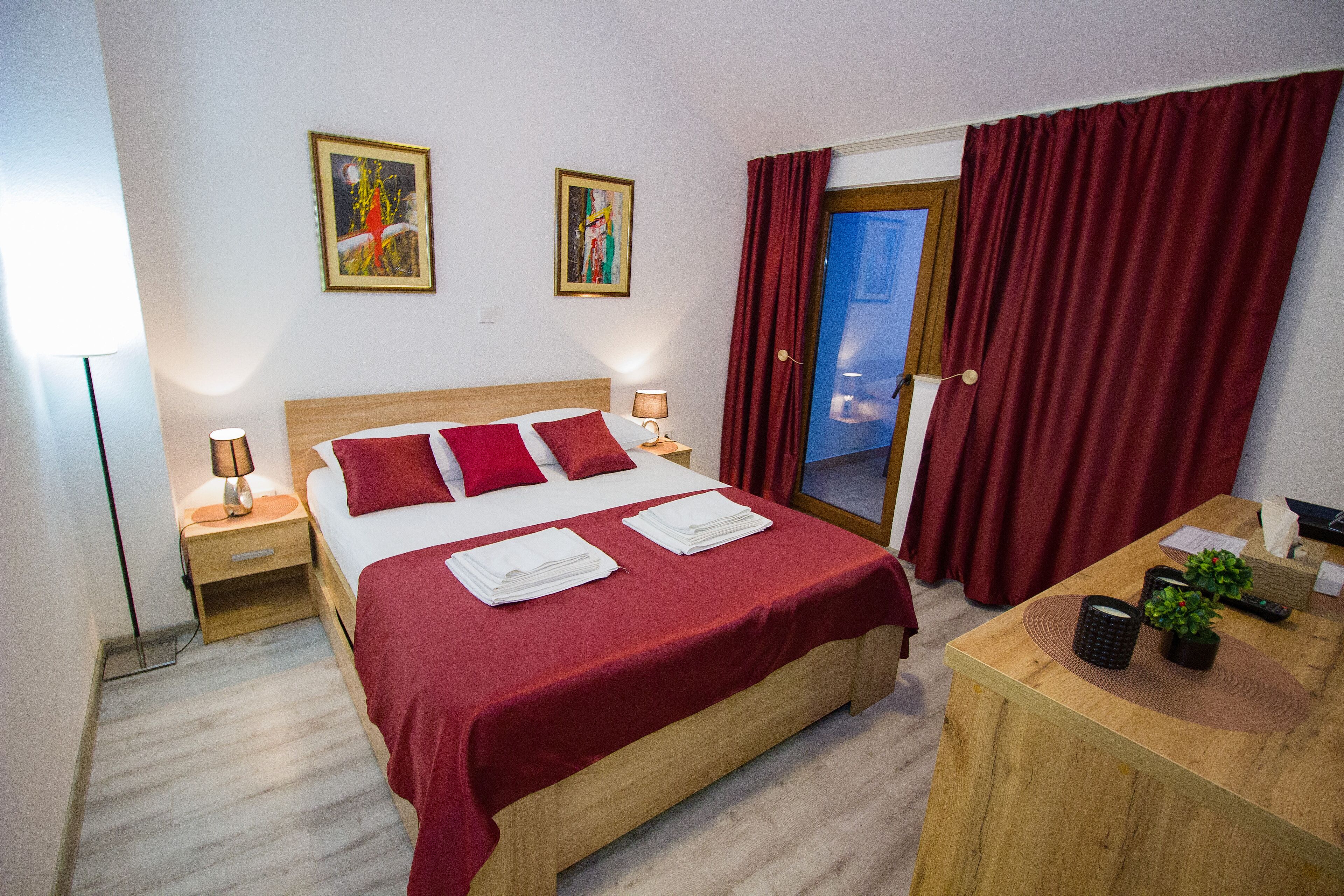 A hotel room with a bed, two bedside tables, a desk, and a window with red curtains.