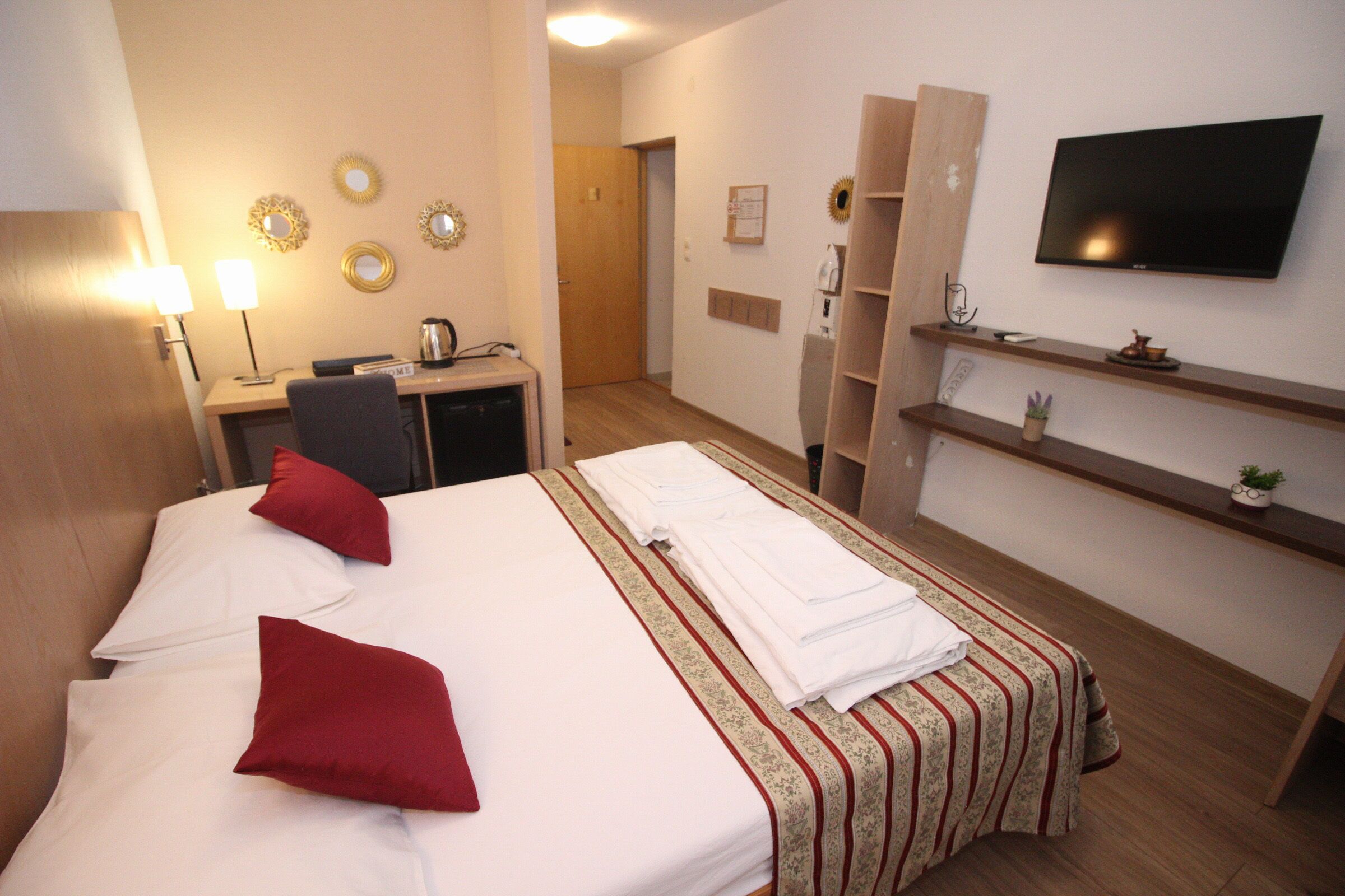 A hotel room with two beds, a desk with a computer, a TV, and a small shelf with a plant.