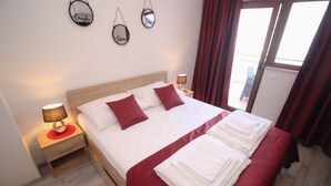 A neatly made bed with red bedding and white pillows, a wooden nightstand with a lamp, and a view of the outdoors through a window with red curtains. - "VILLA DENY" Boutique HOTEL - with free parking (Mostar)