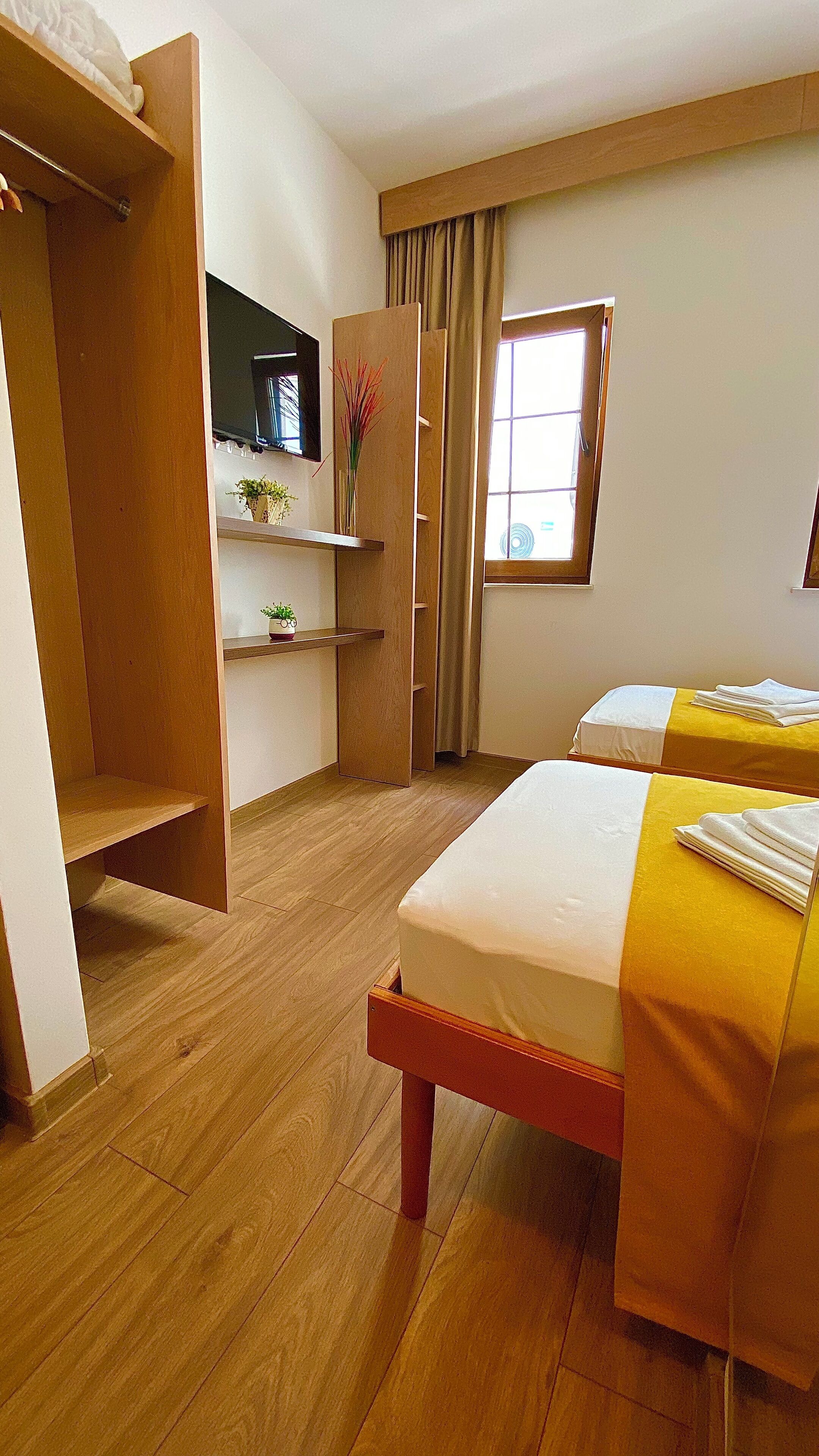 A hotel room with two beds, wooden flooring, a wardrobe, a TV, and a window with curtains.