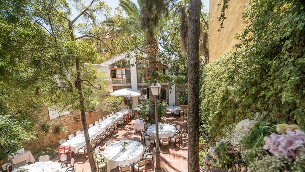An outdoor dining area with round tables and chairs, surrounded by greenery and a building in the background. - Hotel Nou Romà (Denia)
