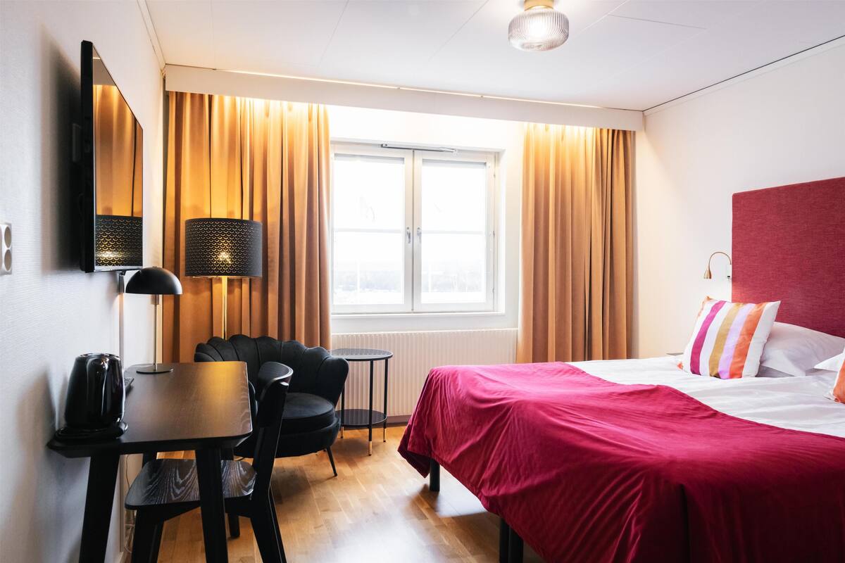 A hotel room with a bed, a desk with a chair, a lamp, a television, and a window with curtains.