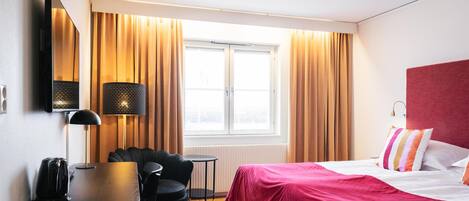 Habitación de hotel con cama, escritorio con silla, lámpara, televisor y ventana con cortinas.
