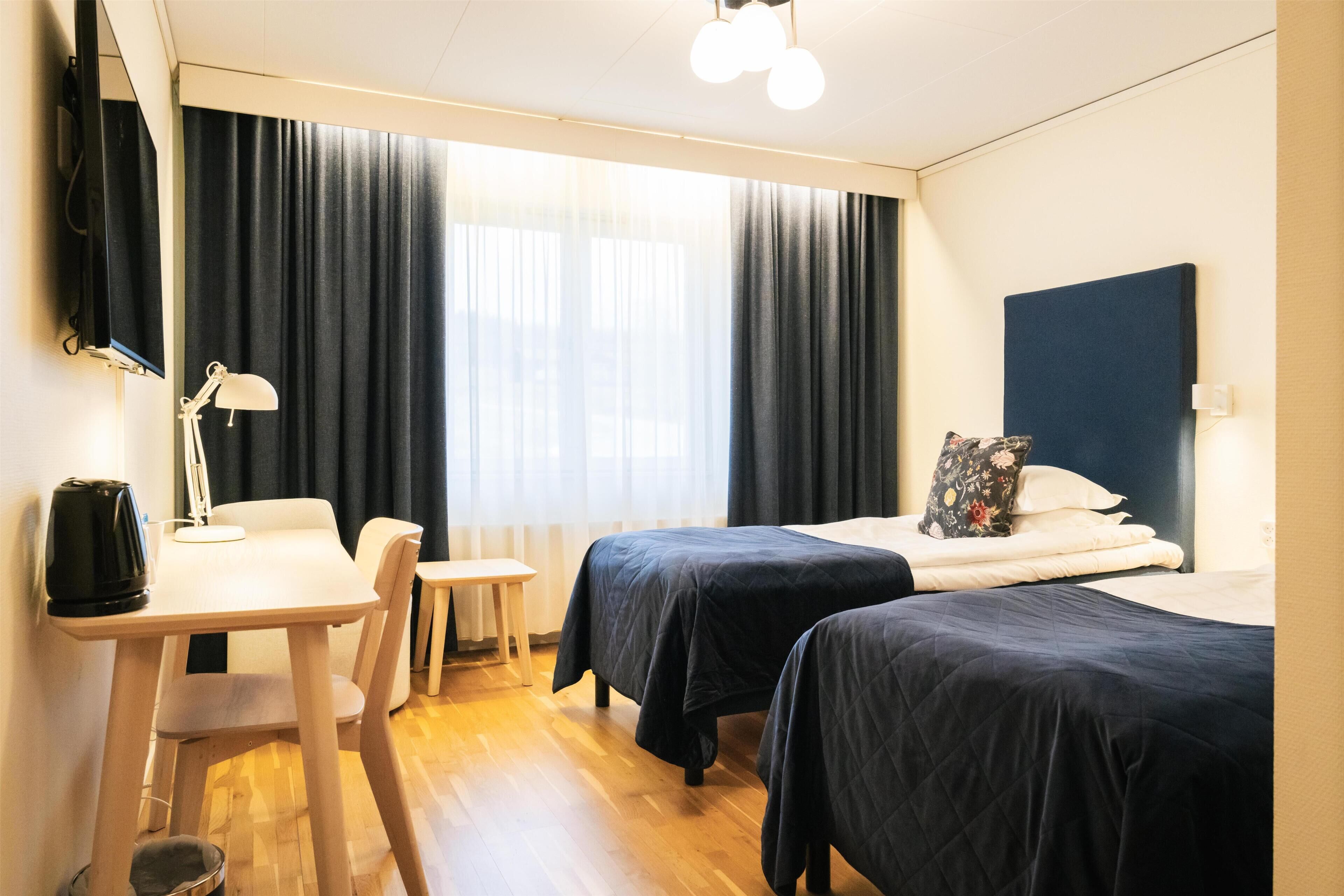 Habitació d'hotel amb dos llits, una taula de treball, una cadira, una làmpada i una finestra gran amb cortines.