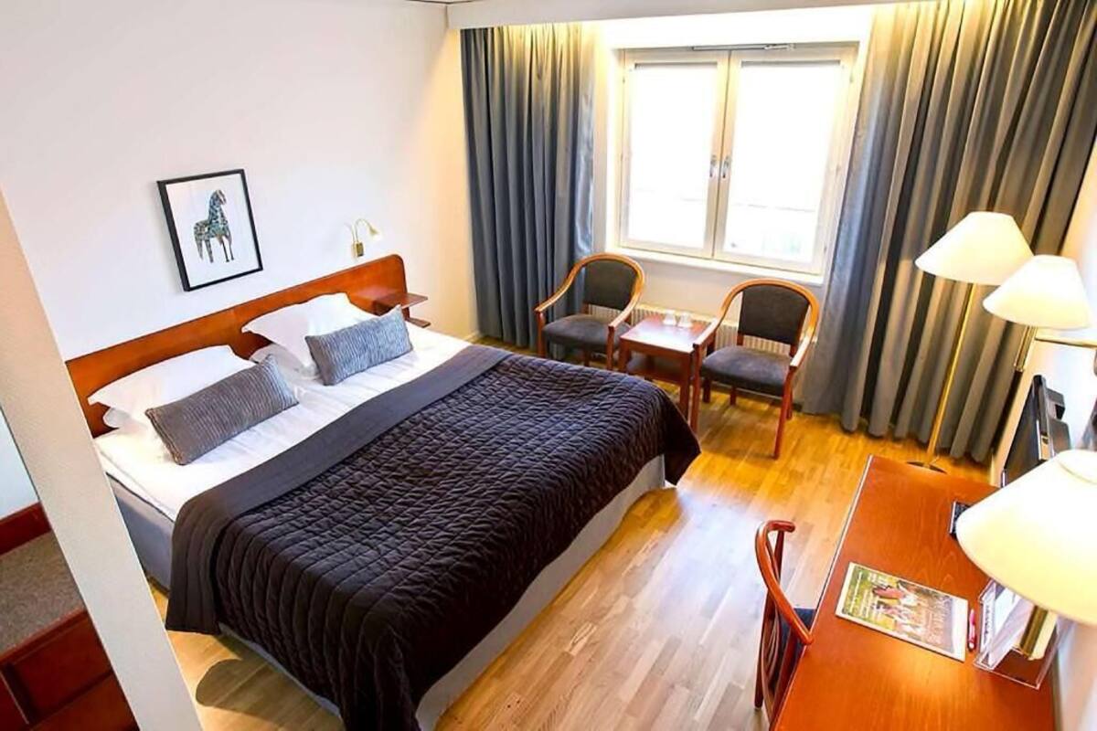 A hotel room with a bed, a desk, two chairs, a window with curtains, and a framed picture on the wall.