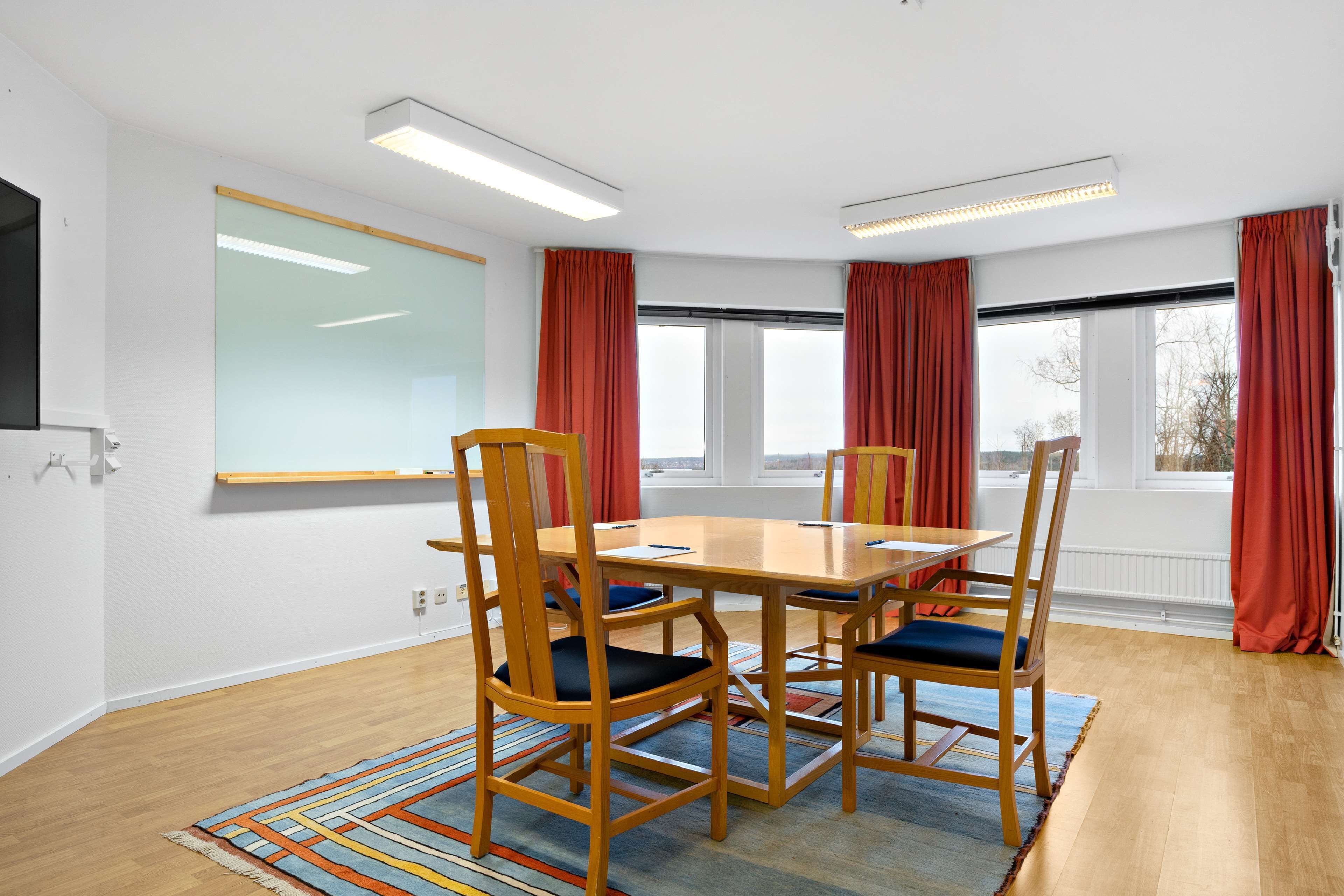 A conference room with a wooden table, four chairs, a whiteboard, and large windows with red curtains.