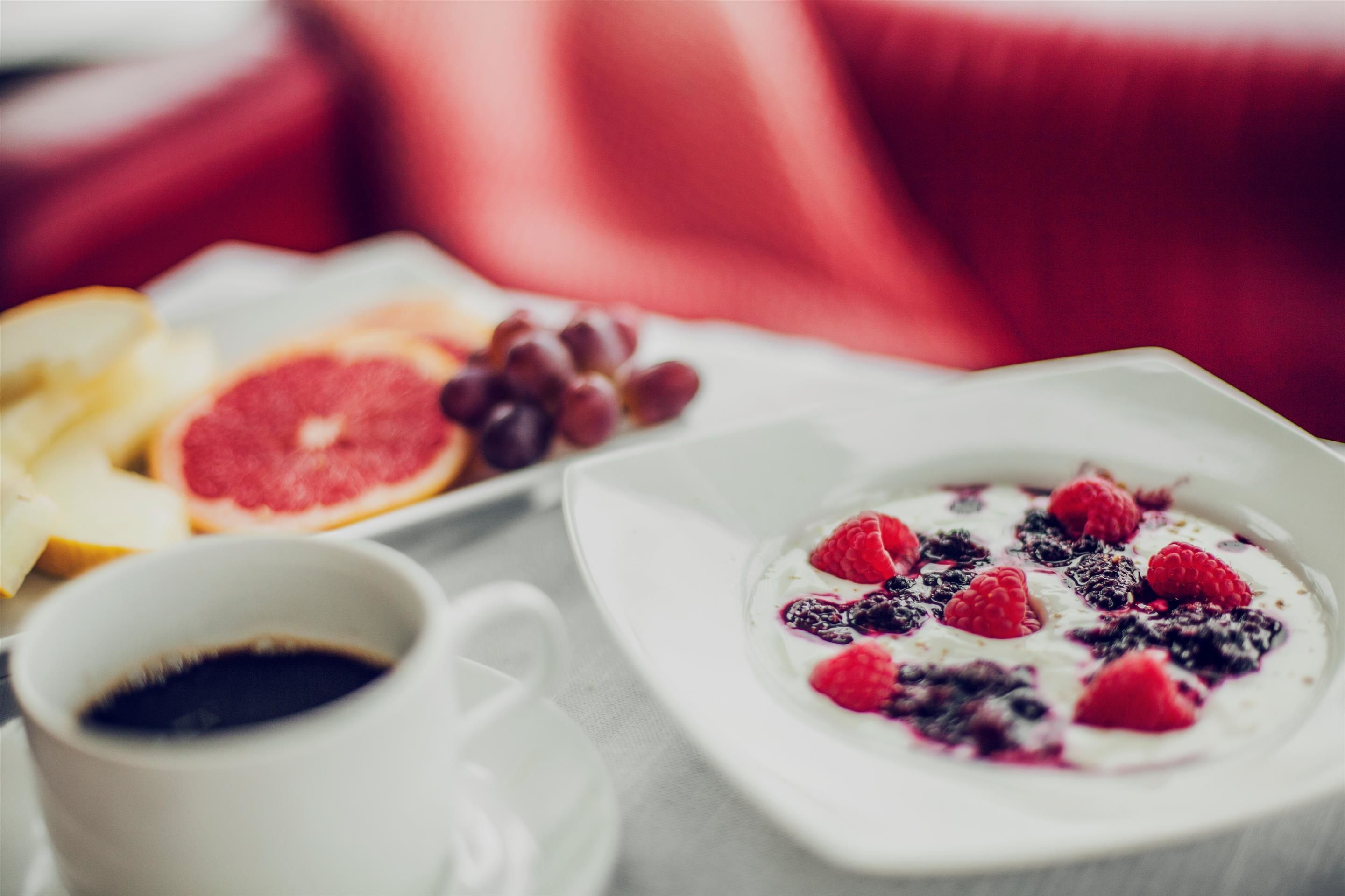 Un desayuno con un plato blanco que contiene yogur cubierto de frutos rojos, una taza de café y fruta cortada.